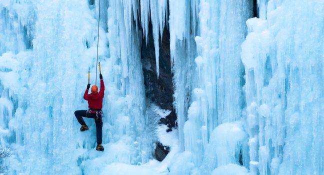 Athletic man in red coat with orange helmet and gold ice tools climbing a large blue wall of ice at the Ouray Ice Park