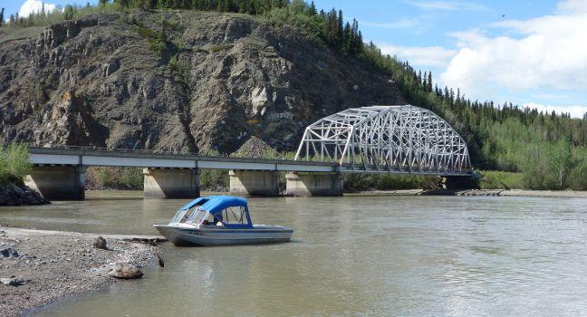 A scenic picture of a bridge in alaska with a small boat on a river.